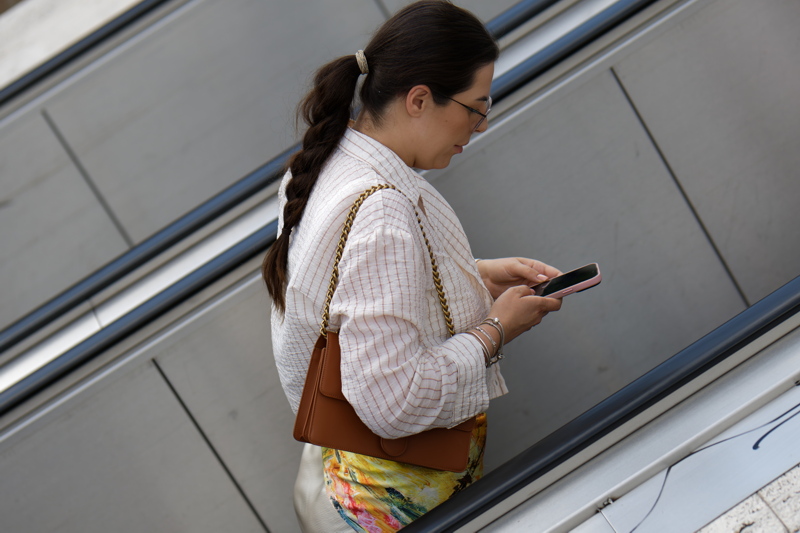 27 - Woman looking down at her phone while wearing a cream-colored, red-striped poplin blouse with pearl details from Zara